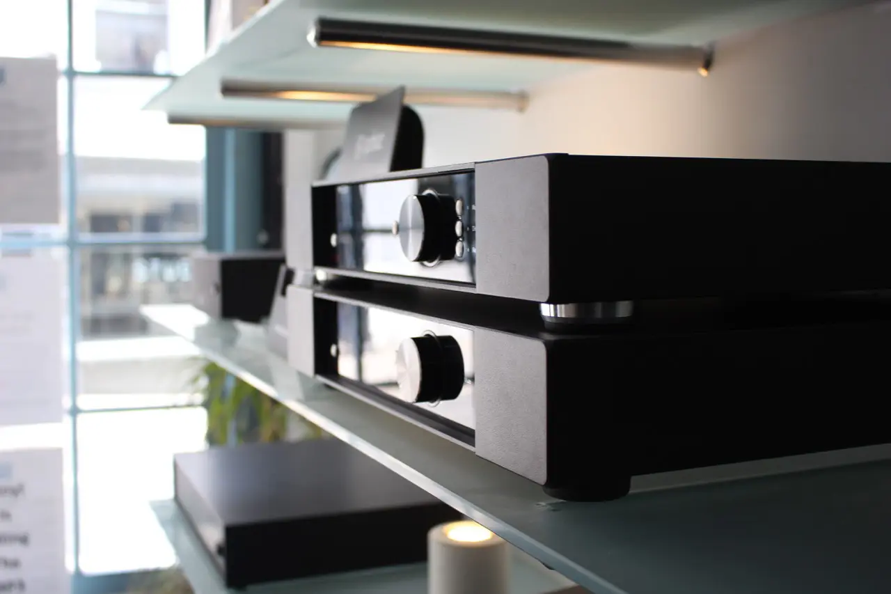 Two stacked black hi-fi audio components with metal control knobs, displayed on a glass shelf under warm lighting in a modern showroom setting