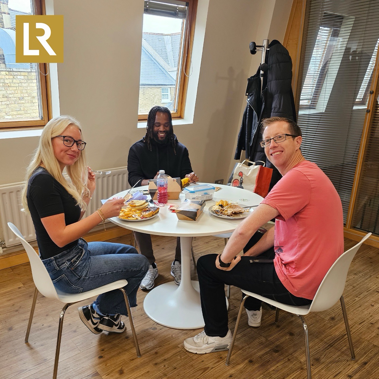 Three colleagues from LR Legal smiling and enjoying a Nando's lunch together at a small round table in a bright office space with wooden floors and large windows